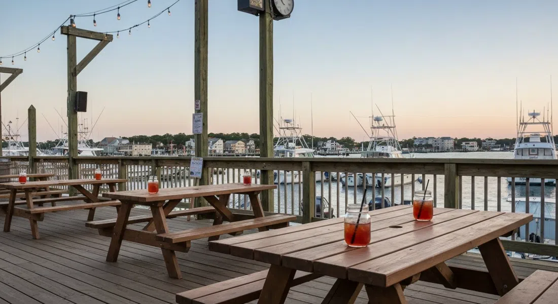 Terraza de Back Bay Ale House en Gardner’s Basin a la luz del atardecer, cócteles en tarros de albañil sobre mesas de picnic con barcos de pesca y agua de la bahía interior al fondo
